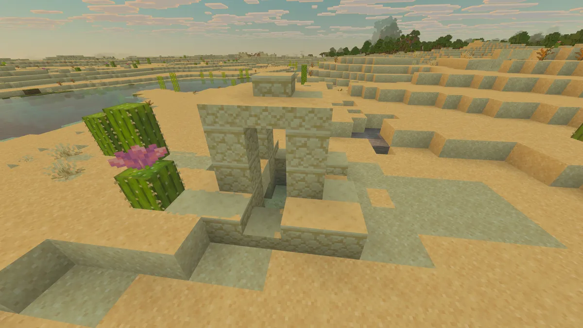 A small sandstone well in a desert with cactus beside it, flat sand all around and a lake and trees in the distance