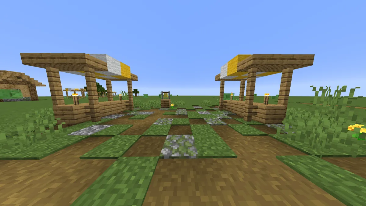 Two wooden market stalls with striped yellow and white awnings facing each other across a path of dirt and grass, with a bell on a post between them and a village house in the distance