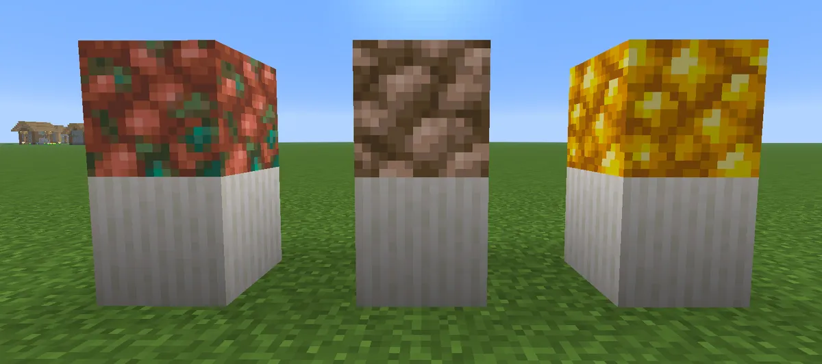 A block of raw copper, a block of raw iron and a block of raw gold, each on top of a white block, standing in a row on a grass field