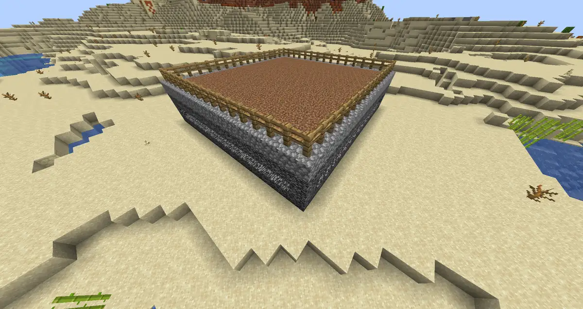 A square spleef arena in a Minecraft desert, with a dirt floor surrounded by an oak fence on top of layers of stone and cobblestone