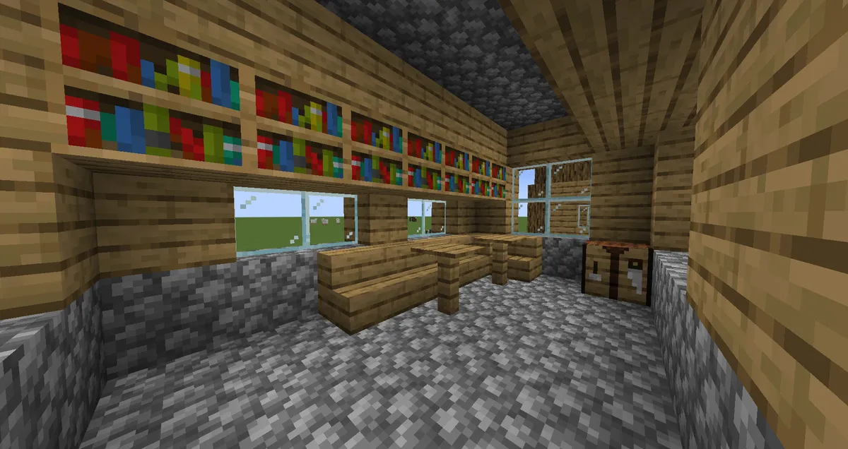 The inside of a library in a Minecraft village, with bookshelves along the top of the wall, oak stairs used as seats at plank tables, glass windows and a crafting table in the corner