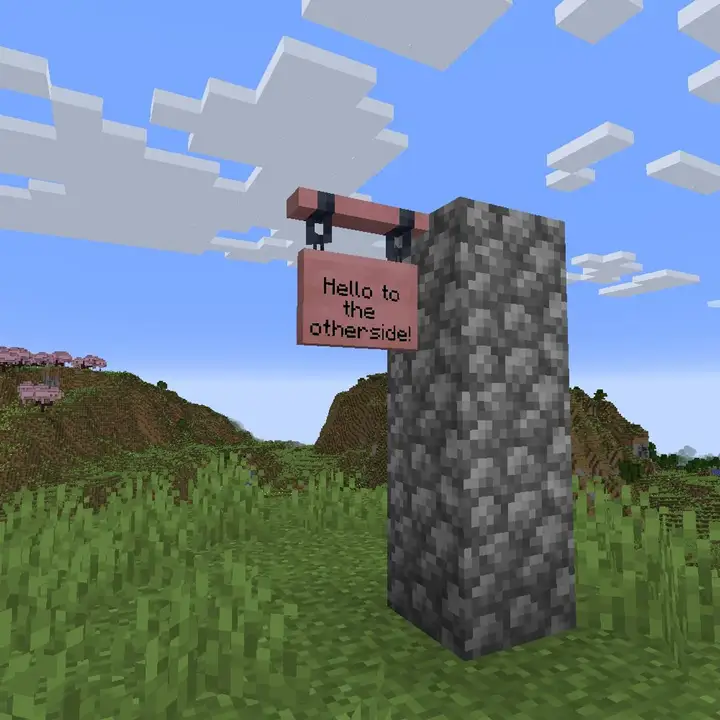 A pink cherry wood hanging sign reading Hello to the otherside! hanging from a cobblestone pillar in a grassy meadow, with a cherry grove on the hills behind it