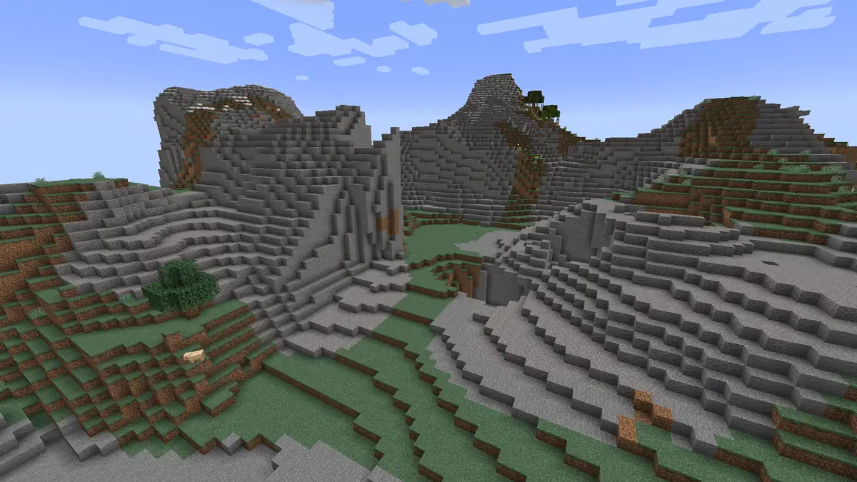 Bare stone hills in a windswept gravelly hills biome with patches of grass and dirt, a few small trees and steep untouched slopes under a blue sky with blocky clouds