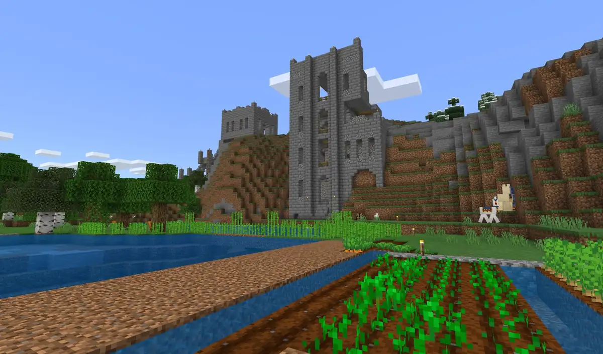 A ruined stone brick castle with a tall tower built into a hillside, above a lake, a dirt path along the water and a field of crops, with llamas grazing below the walls