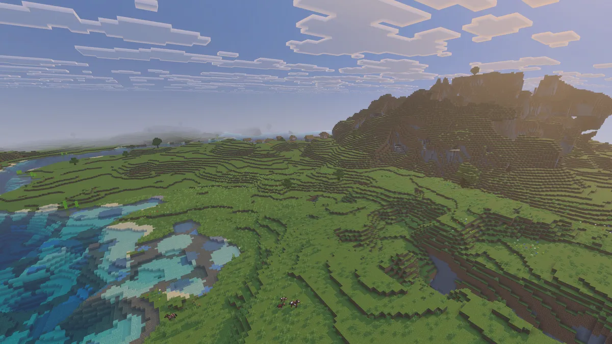 Wide green plains seen from above, with a shallow lake on the left, a few animals in the grass and a rocky hill rising on the right
